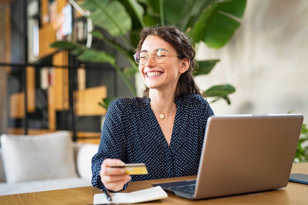 Young happy woman doing shopping online with her laptop at home. Portrait of excited woman holding credit card and buy on an e-commerce site with copy space. Beautiful laughing girl paying online bills using debit card and computer.